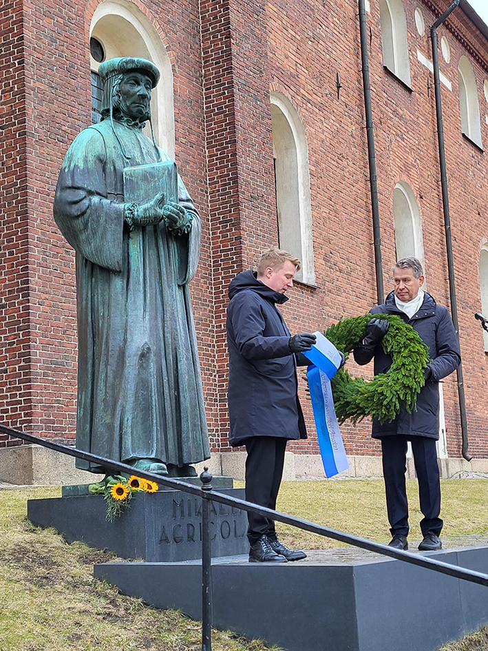 Agricolan patsas Tuomiokirkon äärellä, kaksi henkilöä ja seppele, jossa sinivalkoiset nauhat.