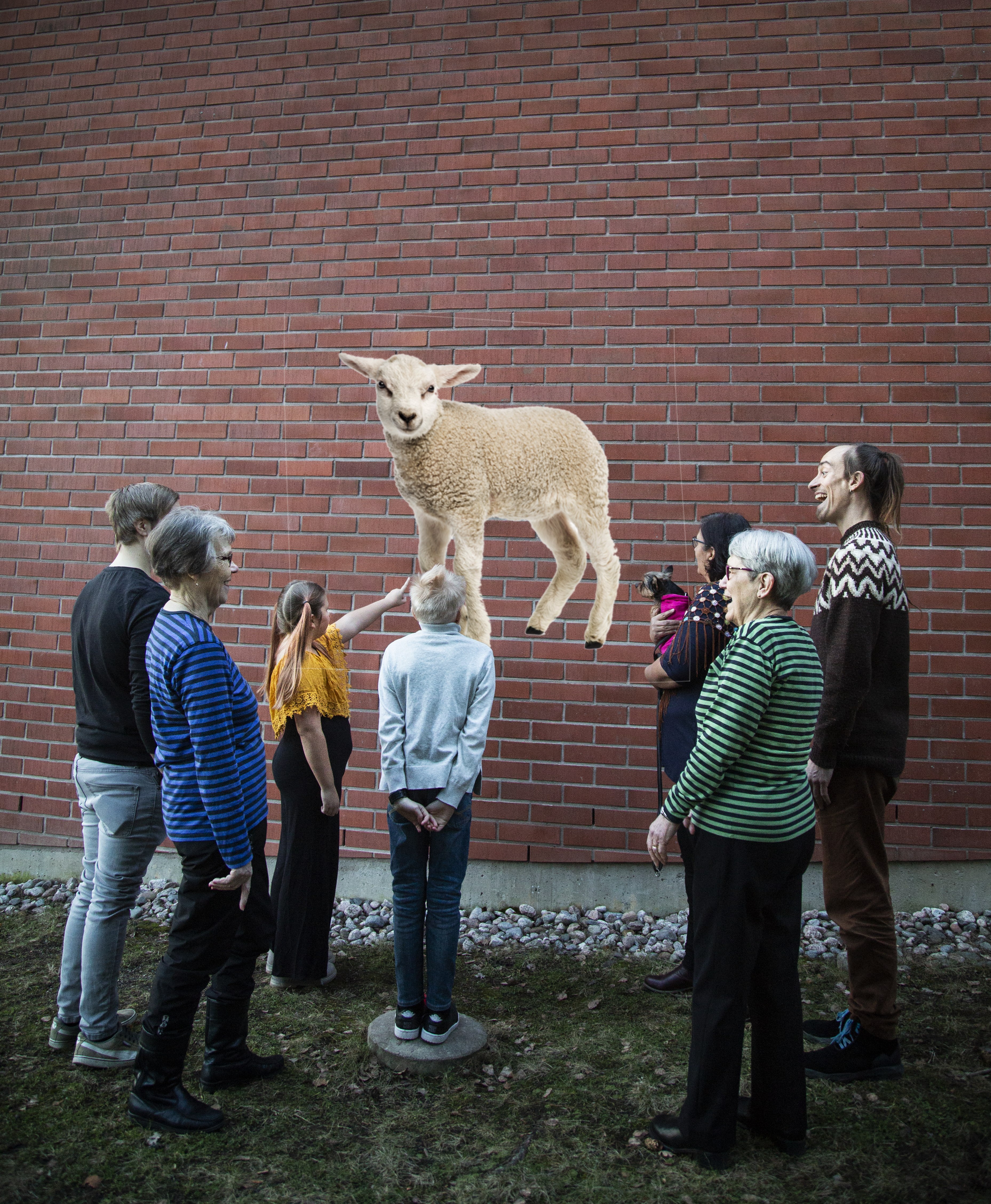 Tiiliseinä edessä eri ikäisiä ihmisiä, jotka tuijottavat seinässä olevaa karitsan kuvaa