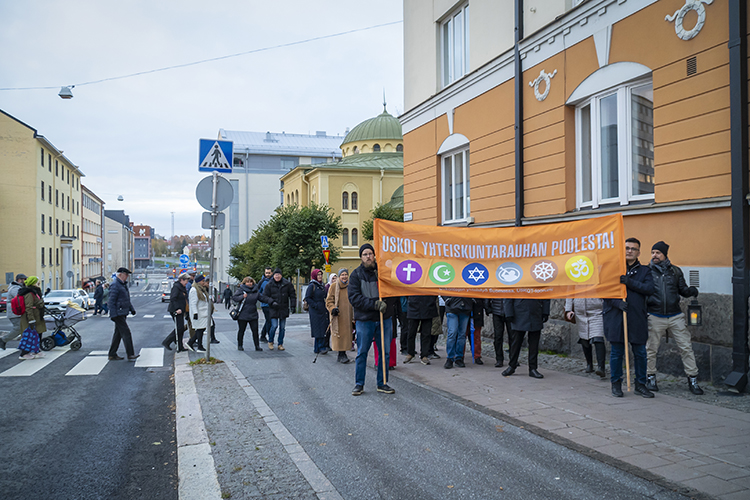 Iso joukko ihmisiä kantaa kadulla kulkueessa suurta banneria, jossa on eri uskontojen symboleita ja teksti ...