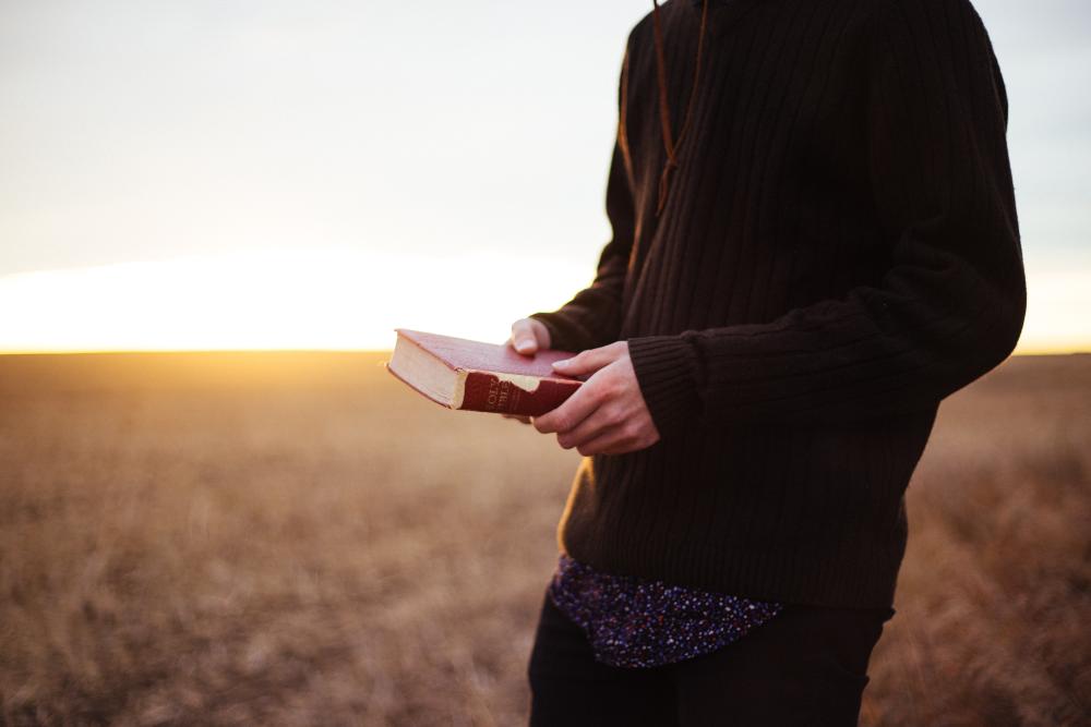 Tummiin vaatteisiin pukeutunut henkilö pitää kädessään Raamattua. Taustalla pelto ja auringonlasku.