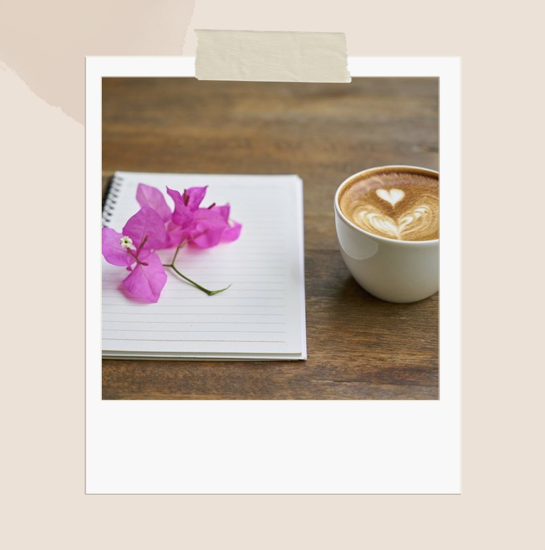 Puupöydällä maitokahvi ja vihko, jonka tyhjällä sivulla pieni punainen kukka.