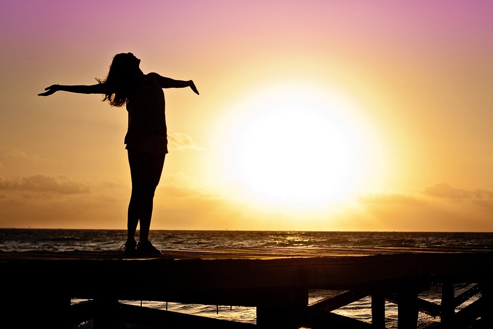Naishenkilö seisoo laiturilla käsivarret vapautuneesti sivuille levitettyinä. Taustalla meri ja auringonlasku.