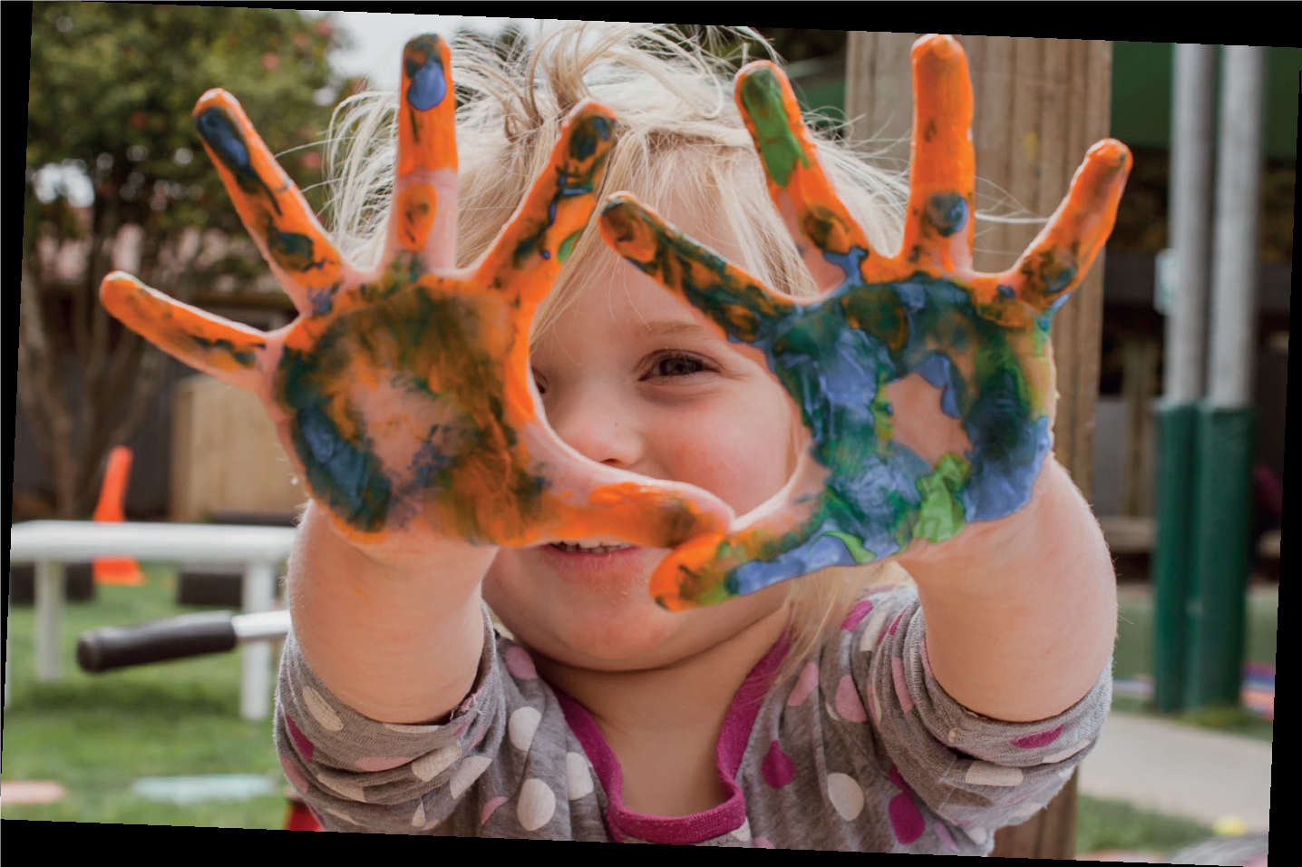 Lapset kädet ovat täynnä erivärisiä maaliroiskeita. Tyttö kurkistaa käsien takaa.