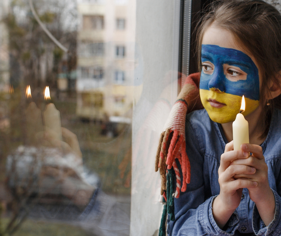 Tyttö, jolla on kasvomaalattu ukrainan lippu katsoo ulos ikkunasta kynttilä kädessä.