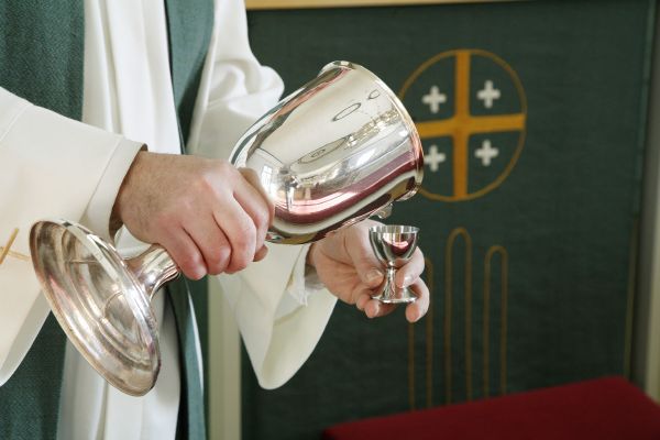 Kuvassa pappi jumalanpalvelusvaatteissa kaataa ehtoollisviiniä maljasta pikariin.