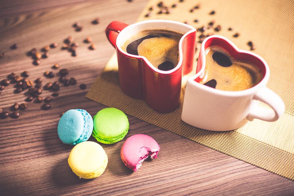 Kaksi sydämen muotoista kahvikuppia sekä värikkäitä macarons-leivoksia puupöydällä. Pöydällä kahvipapuja sekä pöytäliina.
