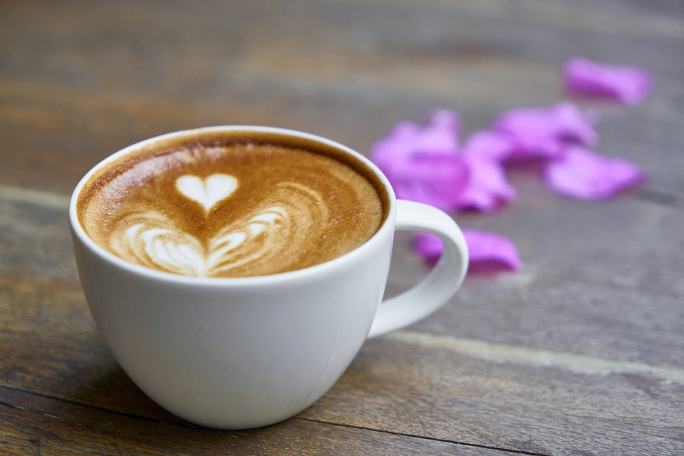 Kahvikuppi, jossa sydän tehtynä kahvin vaahdolla. Kahvikuppi on puupöydällä ja sen takana on kukan terälehtiä.