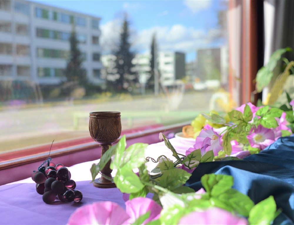 Pääsiäisikkunat Maarian seurakunnassa Kuvassa näkyy ehtoollispikari ja kukkia ja ikkunan takana naapurustoa.