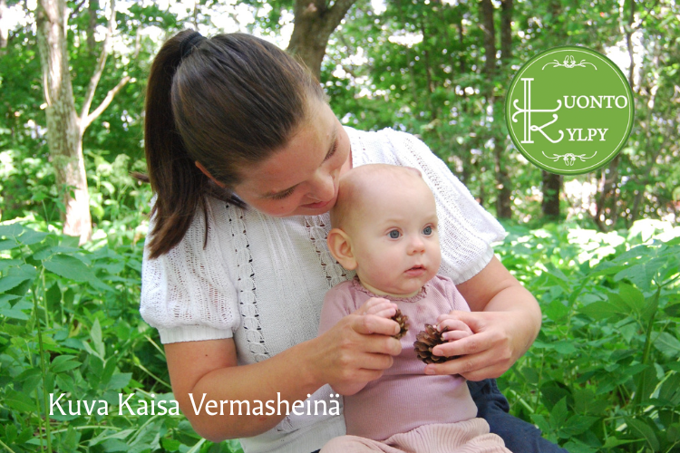 Vauvojen luontokylpyryhmä Kuvassa vauva istuu metsässä äidin sylissä kädessään käpy ja tekstissä sanotaan kuva Kaisa Vermasheinä.