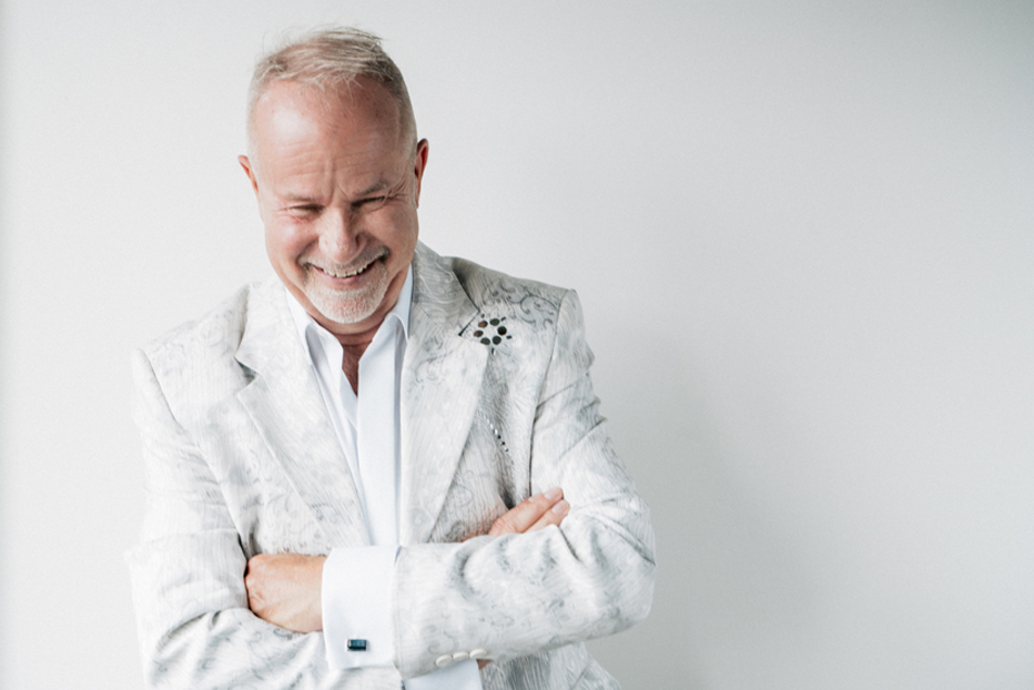 Kuvassa laulaja Petri Laaksonen hymyilee valkeassa asussa ja valkeassa taustassa ja katsoo alaspäin.
