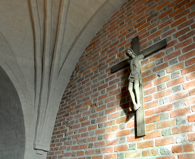 Pappi tavattavissa klo 13-17 Tiiliseinää, jolla roikkuu iso vanhannäköinen puinen krusifiksi. Krusifiksin Jeesus katsoo kameraa kohti.