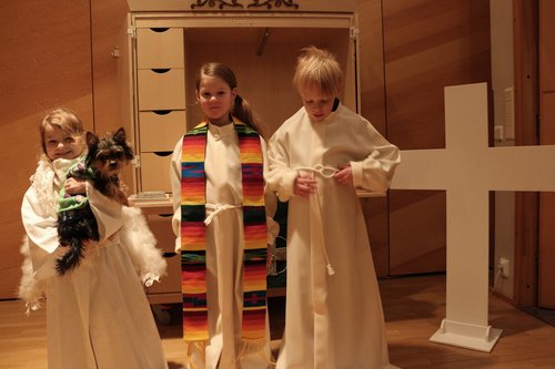 Kolme lasta pukevat päälleen valkeat albat. Yhdellä on sylissä kirkkokoira Muksis. Taustalla näkyy kaappi ja r