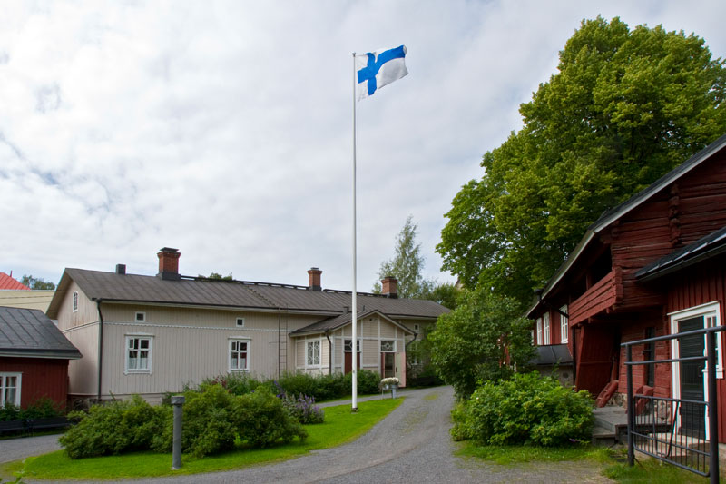 Vaalea, vanha puurakennus, jonka edustalla lipputangossa liehuu Suomen lippu.