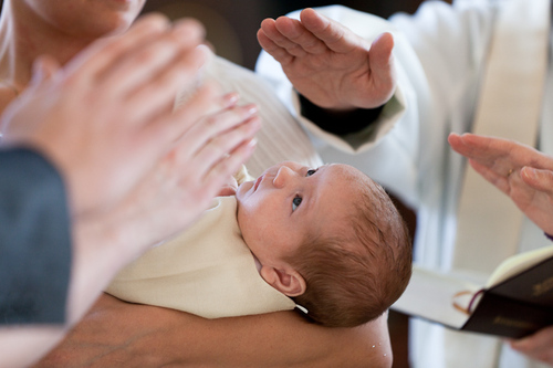 Kuvassa on kastettava lapsi, jonka pään päällä ovat papin, kummien ja vanhempien siunaavat kädet.