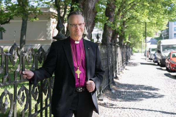 Arkkipiispa seisoo ulkona rauta-aidan vieressä piispan purppuranpunaisessa virkapaidassa, katsoo kameraan ja hymyilee