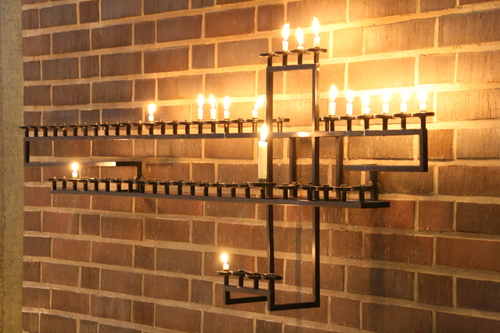 Seinällä ristinmuotoinen kynttilänjalka, jossa paikat usealle kynttilälle. Monta kynttilää palamassa.