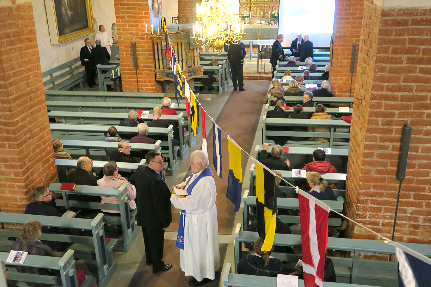 Maarian kirkko sisältä, ihmisiä istuu penkeissä, heidän yllään värikkäitä laivalippuja