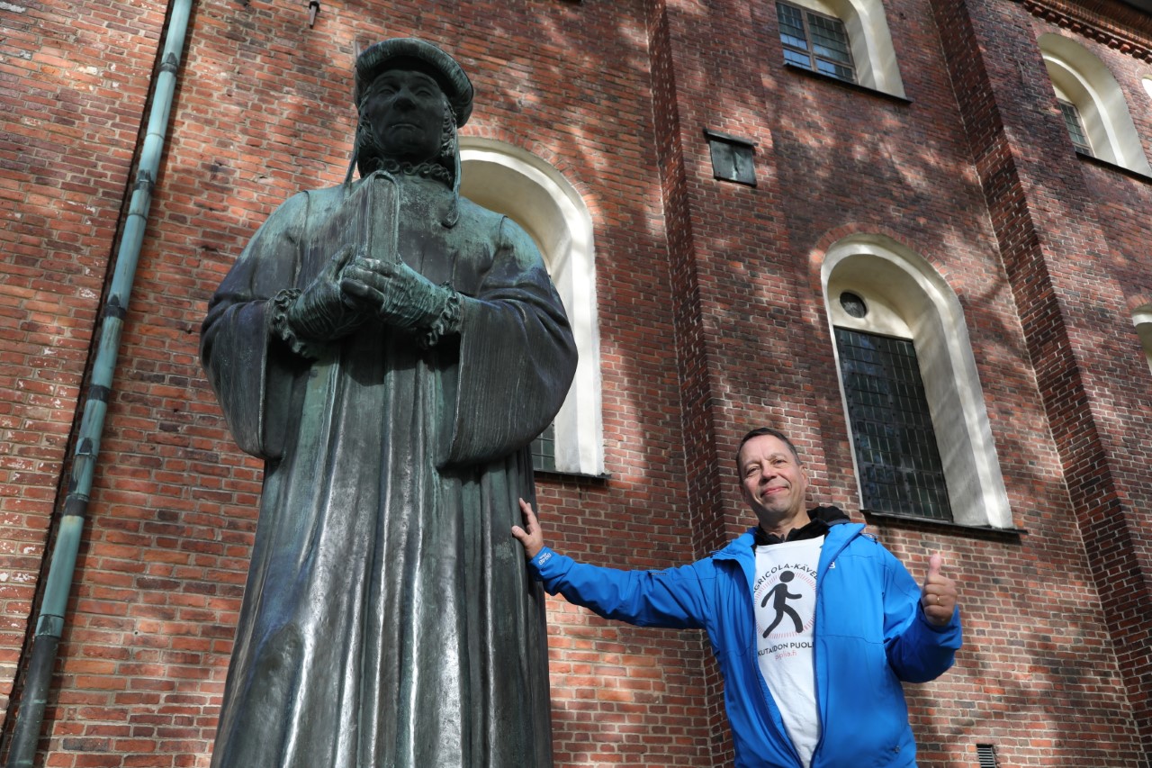Laulutaiteilija Hannu Holma nojaa Tuomiokirkon edellä olevaan Agricola-patsaaseen ja näyttää peukkua.
