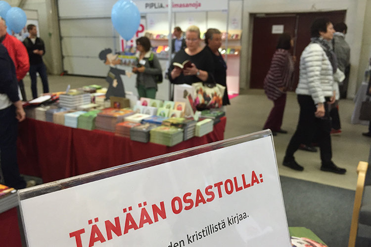 Esittelyosasto Kirjamessuilla, etualalla osittain näkyvissä kyltti messuosaston ohjelmasta, taka-alalla ihm...