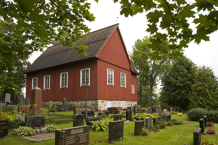 Punainen pieni puukirkko vehreän hautausmaan keskellä.