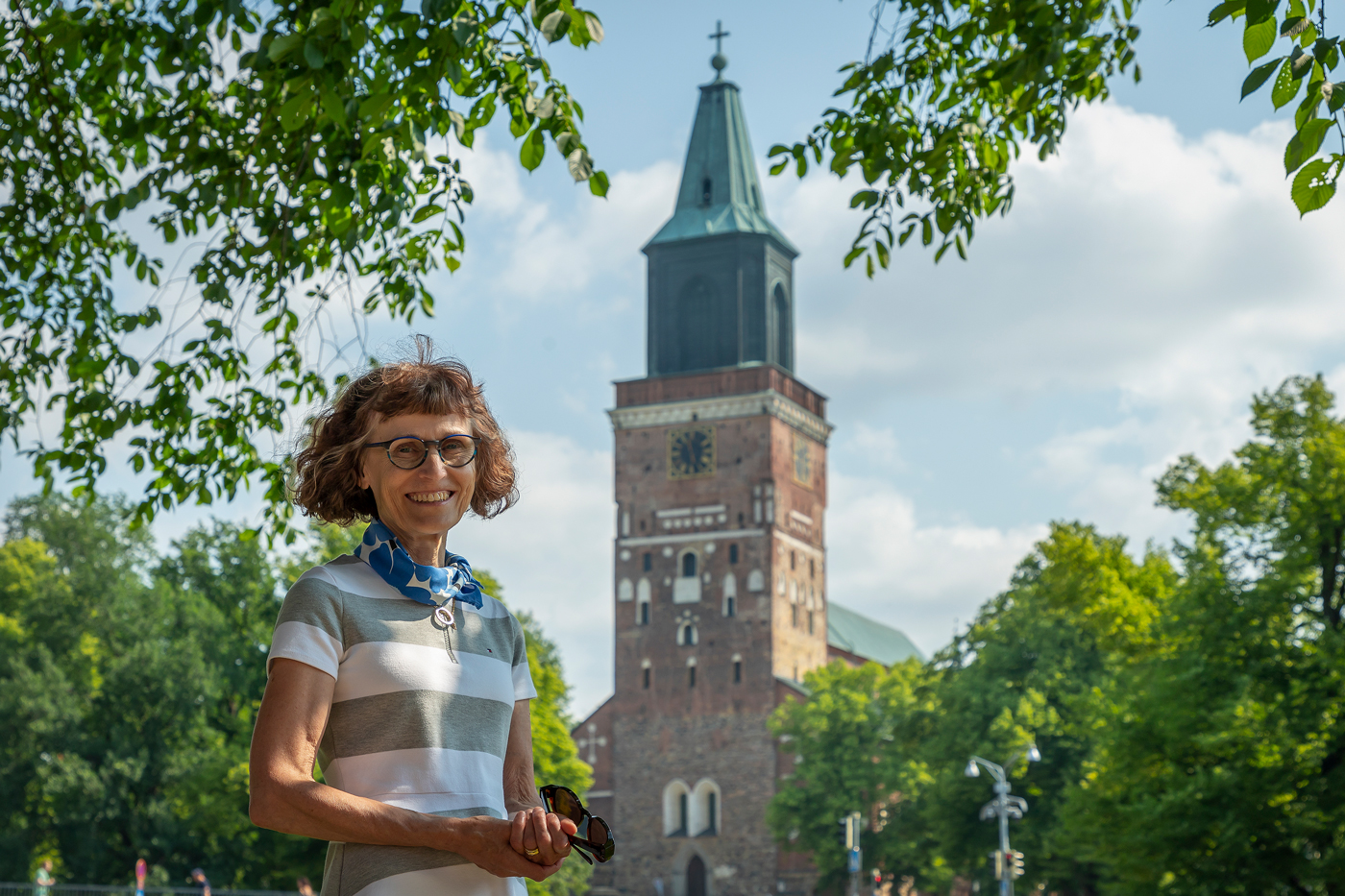 Raidalliseen t-paitaan pukeutunut nainen katsoo hymyillen kameraan kesäisessä maisemassa. Taustalla Turun t...