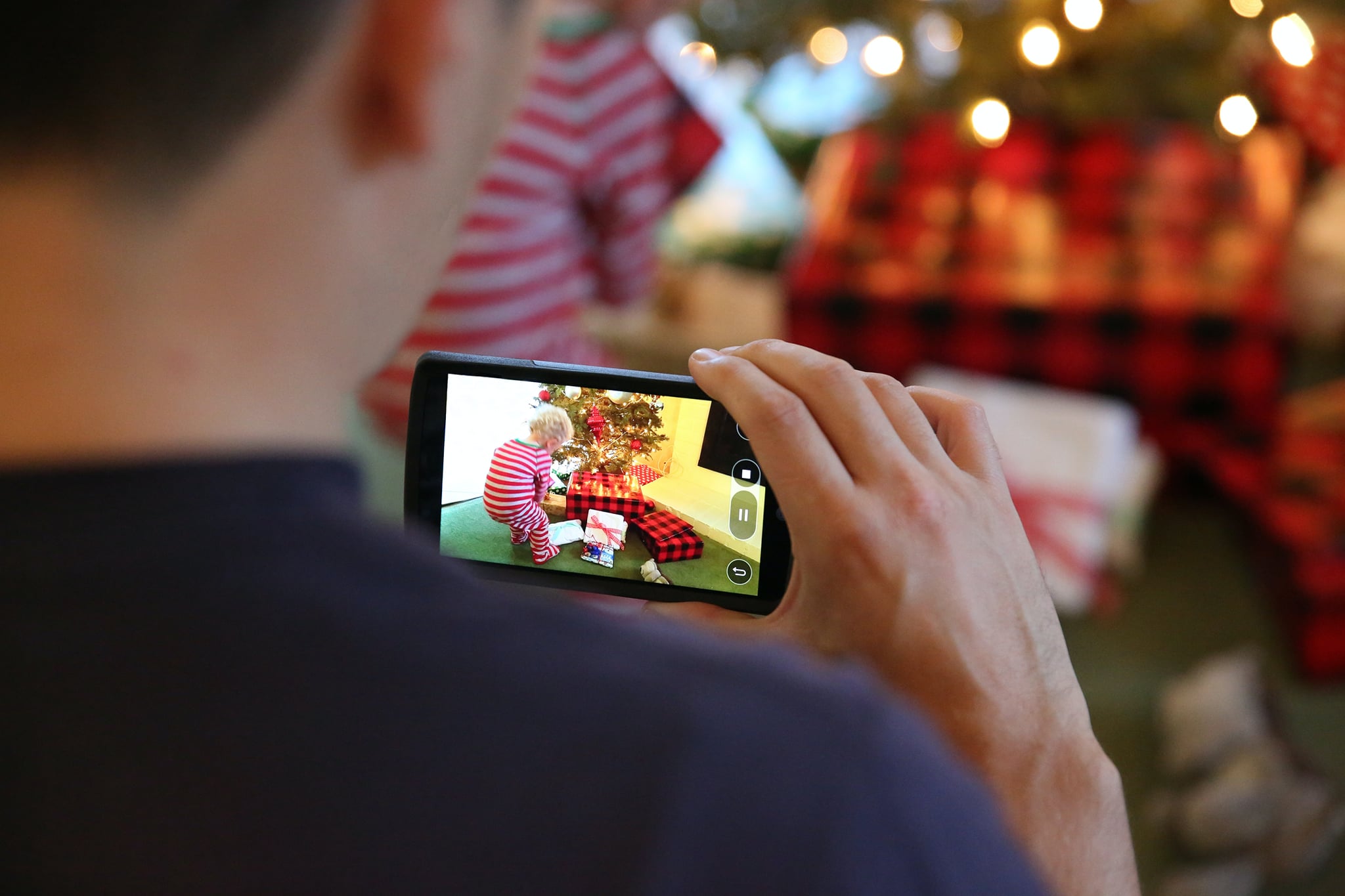Miespuolinen henkilö kuvaamassa kännykällään joulukuusen edessä leikkivää lasta. Kuvituskuva.