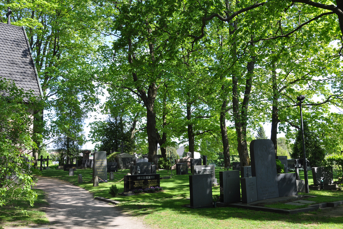 Tummia hautakiviä, hiekkakäytävä ja puita kesäisen vehreällä hautausmaalla.